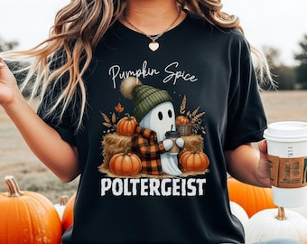 Op de afbeelding: Een zwart t-shirt met korte mouwen en een afbeelding van een geest met een groene muts en een geruit overhemd, die een koffiebeker vasthoudt. De geest is omringd door pompoenen en hooibalen. De tekst luidt Pumpkin Spice POLTERGEIST.
