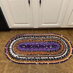 May include: Oval Halloween rug with a purple centre and concentric bands of orange, black, white, and purple fabrics. The rug is placed on a tiled floor in front of white cabinets.