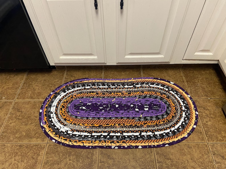 May include: Oval Halloween rug with a purple centre and concentric bands of orange, black, white, and purple fabrics. The rug is placed on a tiled floor in front of white cabinets.