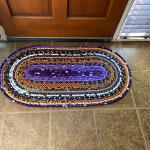 May include: Oval braided rug with a purple centre and alternating bands of orange, black, white, and patterned fabrics. The rug is placed on a tiled floor near a wooden door.