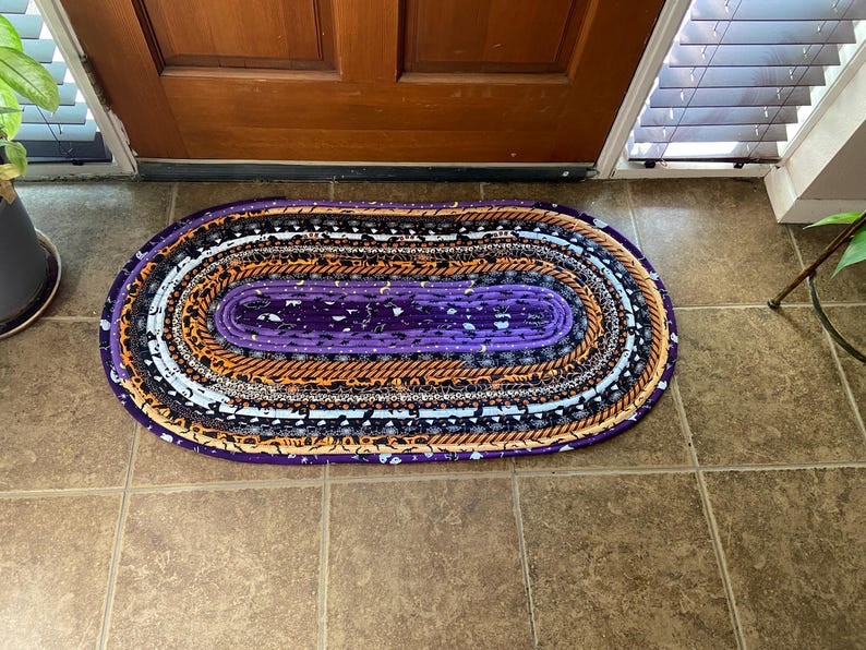 May include: Oval braided rug with a purple centre and alternating bands of orange, black, white, and patterned fabrics. The rug is placed on a tiled floor near a wooden door.