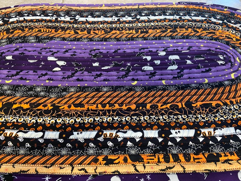 May include: Oval Halloween-themed rug made from fabric strips. The rug has orange, black, and purple stripes with Halloween patterns such as bats, ghosts, and spiderwebs. A decorative item for the home.