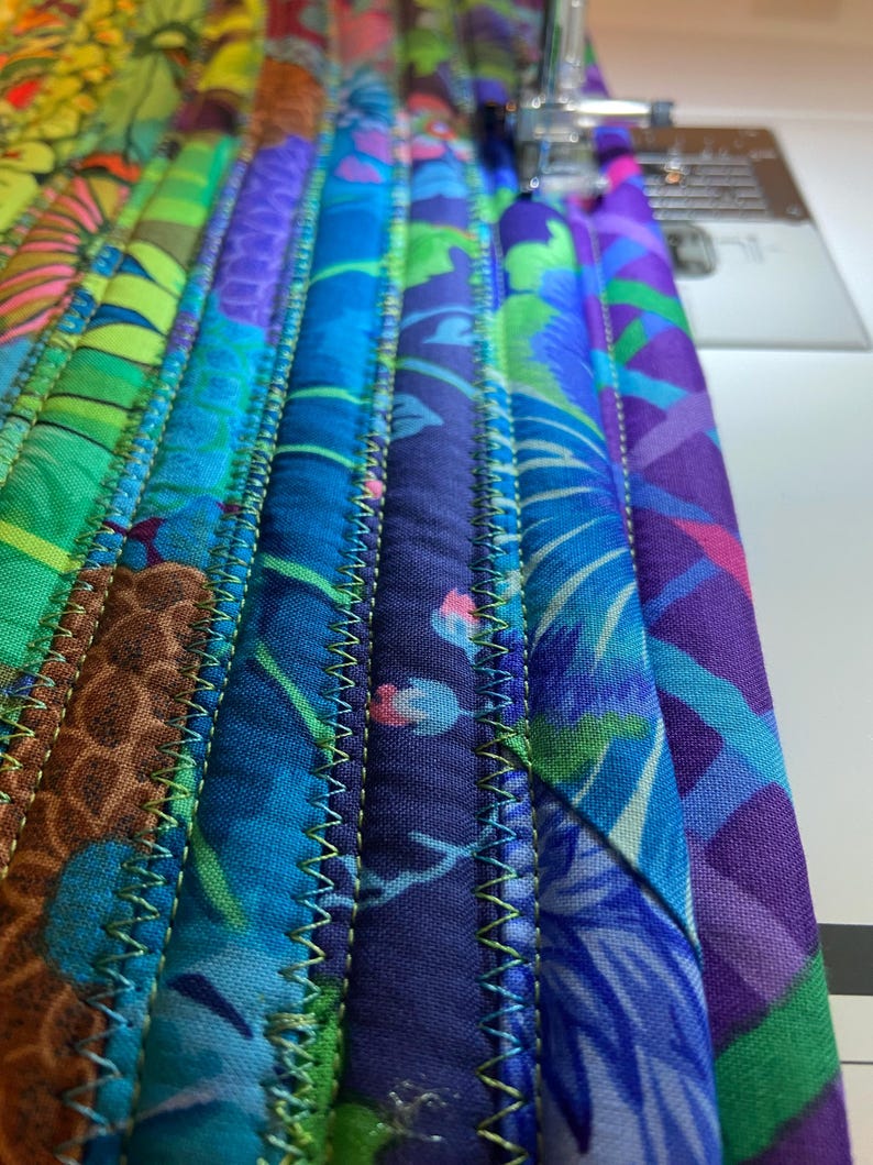 Op de afbeelding: Close-up van een kleurrijke gewatteerde stof in tinten blauw, groen, paars en bruin. De stof is gestikt met een zigzagpatroon met groen garen. De naald van de naaimachine is zichtbaar in de rechterbovenhoek.