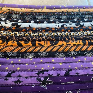 May include: A Halloween-themed fabric item with horizontal stripes in black, orange, and purple. The stripes feature patterns of bats, spiderwebs, cats, and stars. The item is likely a table runner or placemat.