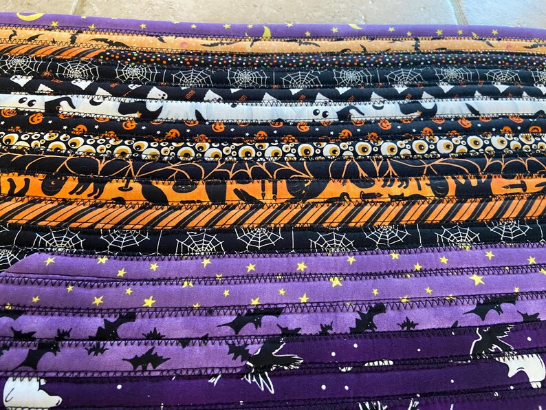 May include: A Halloween-themed fabric item with horizontal stripes in black, orange, and purple. The stripes feature patterns of bats, spiderwebs, cats, and stars. The item is likely a table runner or placemat.