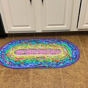 Op de afbeelding: Een ovaal gevlochten vloerkleed met een levendig regenboogkleurenschema. Het kleed heeft een centraal paars gedeelte omgeven door concentrische ringen van groen, geel, blauw en paars. Het kleed ligt op een bruine tegelvloer.