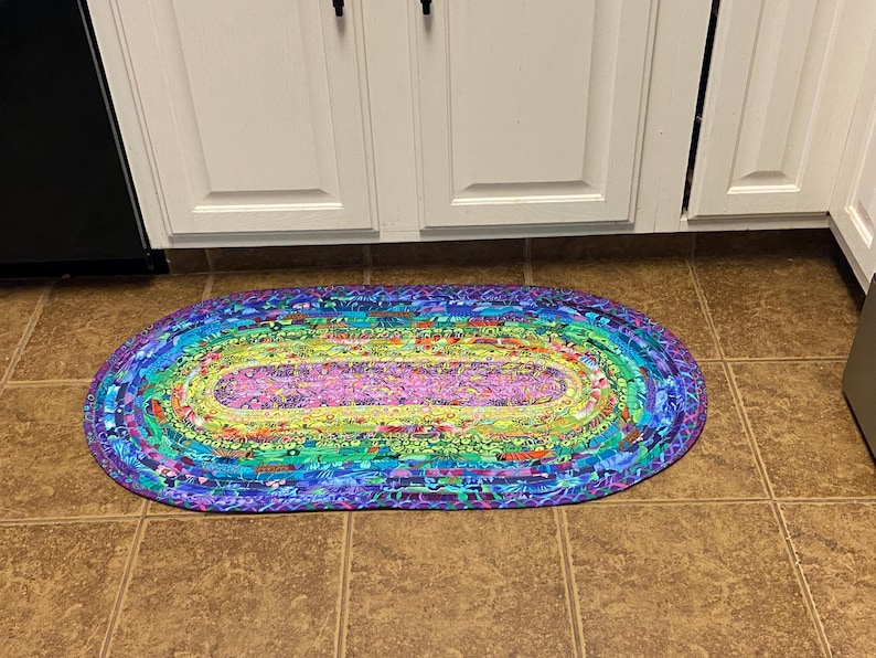 Op de afbeelding: Een ovaal gevlochten vloerkleed met een levendig regenboogkleurenschema. Het kleed heeft een centraal paars gedeelte omgeven door concentrische ringen van groen, geel, blauw en paars. Het kleed ligt op een bruine tegelvloer.
