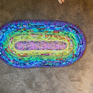Op de afbeelding: Een ovaal gevlochten tapijt met een levendig, veelkleurig ontwerp. Het tapijt heeft een spectrum aan kleuren, waaronder blauw, groen, paars en geel. De gevlochten constructie voegt textuur en visuele interesse toe.