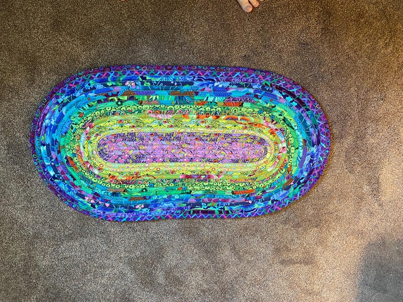 Op de afbeelding: Een ovaal gevlochten tapijt met een levendig, veelkleurig ontwerp. Het tapijt heeft een spectrum aan kleuren, waaronder blauw, groen, paars en geel. De gevlochten constructie voegt textuur en visuele interesse toe.