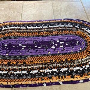 May include: Oval Halloween rug with a purple central section decorated with bats and ghosts. The rug features concentric bands of orange, black, and white patterns, including spiderwebs and stripes. The rug is made of fabric and is designed for indoor use.