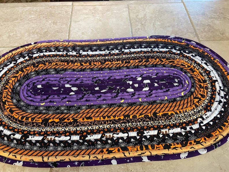 May include: Oval Halloween rug with a purple central section decorated with bats and ghosts. The rug features concentric bands of orange, black, and white patterns, including spiderwebs and stripes. The rug is made of fabric and is designed for indoor use.