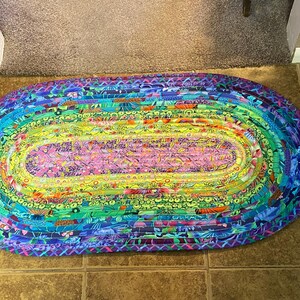 Op de afbeelding: Ovaal gevlochten vloerkleed met een regenboog aan kleuren. Het kleed heeft een roze en gele middensectie, omringd door groene, blauwe en paarse lagen. Het gevlochten ontwerp voegt textuur en visuele interesse toe aan dit woondecoratie-item.