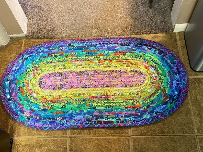 Op de afbeelding: Ovaal gevlochten vloerkleed met een regenboog aan kleuren. Het kleed heeft een roze en gele middensectie, omringd door groene, blauwe en paarse lagen. Het gevlochten ontwerp voegt textuur en visuele interesse toe aan dit woondecoratie-item.