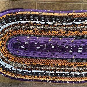 May include: Oval Halloween-themed rug with various patterned fabric strips. The rug features stripes of purple, orange, black, and white fabrics with Halloween motifs like bats, ghosts, and spiderwebs. The rug is approximately 76 cm long.