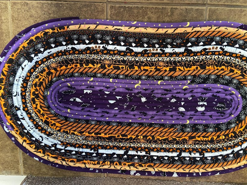 May include: Oval Halloween-themed rug with various patterned fabric strips. The rug features stripes of purple, orange, black, and white fabrics with Halloween motifs like bats, ghosts, and spiderwebs. The rug is approximately 76 cm long.