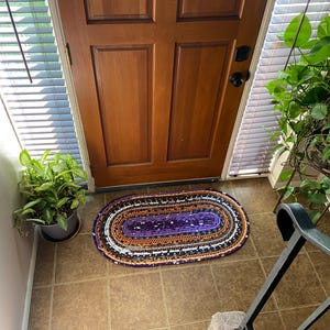 May include: An oval rag rug in purple, orange, black, and white sits on a tiled floor in front of a wooden door. The rug is made of fabric strips and has a braided look. A potted plant is to the left of the rug. The door is made of wood.