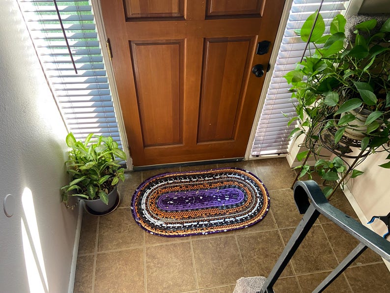 May include: An oval rag rug in purple, orange, black, and white sits on a tiled floor in front of a wooden door. The rug is made of fabric strips and has a braided look. A potted plant is to the left of the rug. The door is made of wood.