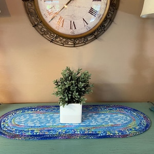 May include: An oval-shaped, blue fabric table runner with a floral pattern. A small, square white planter with a green plant sits on the runner. The runner is approximately 20 inches long.