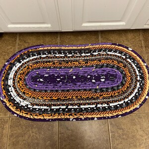 May include: Oval Halloween rug with a purple centre and bands of orange, black, and white fabric. The rug features Halloween patterns like bats and moons. The rug is placed on a brown tiled floor. The rug is approximately 100 cm in length.
