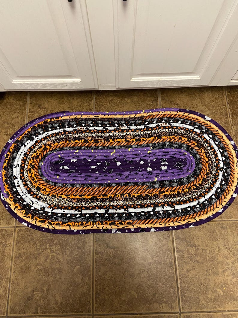May include: Oval Halloween rug with a purple centre and bands of orange, black, and white fabric. The rug features Halloween patterns like bats and moons. The rug is placed on a brown tiled floor. The rug is approximately 100 cm in length.