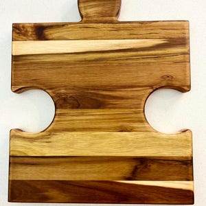 May include: A wooden puzzle-shaped cutting board with a handle. The board is made of various shades of brown wood, with lighter and darker grains creating a natural pattern. The design includes two curved cutouts and a rounded handle.