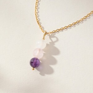 May include: A gold chain necklace featuring a vertical pendant composed of three round beads arranged in a gradient. The top bead is translucent white, the middle bead is pale pink, and the bottom bead is a deep purple amethyst stone.