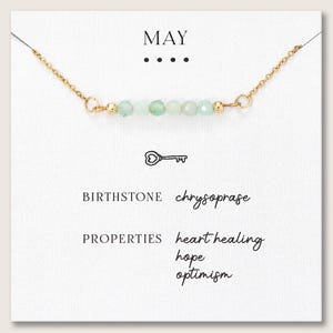 May include: A gold chain necklace with a row of light green beads. The card behind the necklace says "MAY" and "BIRTHSTONE chrysoprase". The card also lists "heart healing, hope, optimism" as properties.