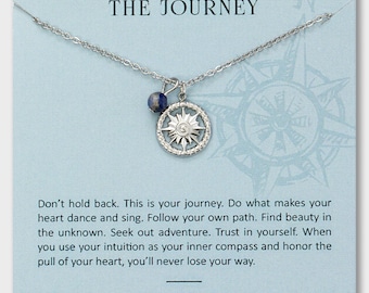 Può includere: Una collana d'argento con un ciondolo a forma di bussola e una perla di sodalite blu, esposta su una carta azzurra. La carta recita "THE JOURNEY" e include un testo ispiratore sulla scoperta di sé e sul seguire il proprio cammino.