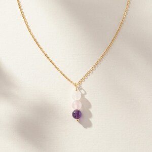 May include: A gold-toned chain necklace featuring a vertical pendant composed of three round beads. The top bead is translucent white, the middle bead is pale pink, and the bottom bead is a deep purple, all suspended from a thin metal link chain.