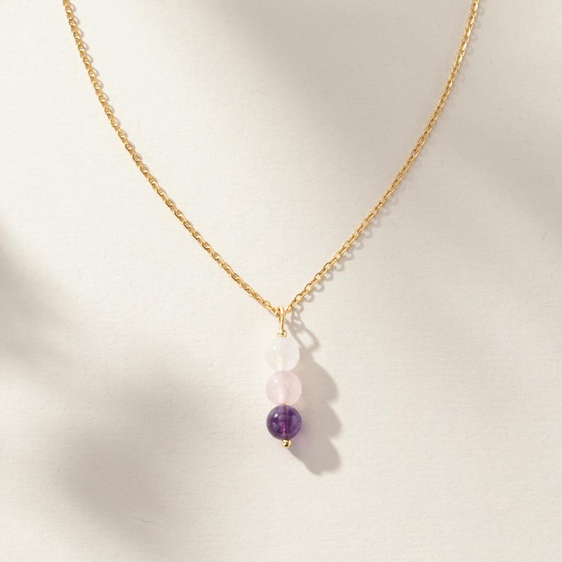 May include: A gold-toned chain necklace featuring a vertical pendant composed of three round beads. The top bead is translucent white, the middle bead is pale pink, and the bottom bead is a deep purple, all suspended from a thin metal link chain.