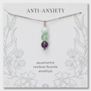 May include: A silver-toned chain necklace featuring a vertical pendant with three round beads: a pale blue aquamarine, a translucent green rainbow fluorite, and a deep purple amethyst. The display card reads ANTI-ANXIETY and lists the stones.