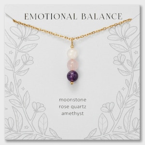 May include: A gold chain necklace featuring a vertical pendant with three round beads: a white moonstone, a pale pink rose quartz, and a deep purple amethyst. The display card reads EMOTIONAL BALANCE, moonstone, rose quartz, and amethyst.