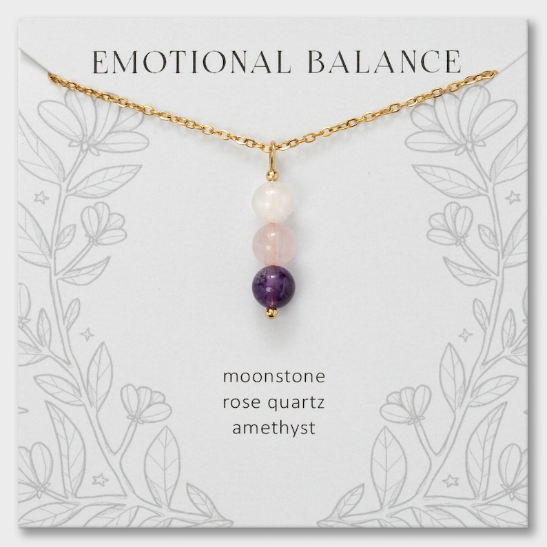 May include: A gold chain necklace featuring a vertical pendant with three round beads: a white moonstone, a pale pink rose quartz, and a deep purple amethyst. The display card reads EMOTIONAL BALANCE, moonstone, rose quartz, and amethyst.