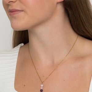 May include: A gold-toned chain necklace featuring a vertical pendant composed of three small, smooth, spherical beads. The beads are arranged in a gradient from a pale cream color at the top to a light pink in the center and a deep purple at the base.