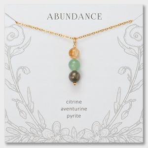 May include: A gold chain necklace with a pendant featuring three stacked spherical beads: a translucent yellow citrine, a light green aventurine, and a metallic pyrite. The display card is printed with the word ABUNDANCE and the stone names.