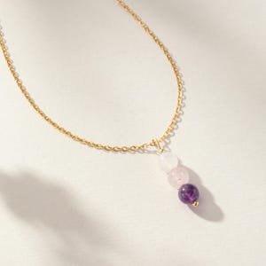 May include: A gold chain necklace featuring a vertical pendant composed of three round beads arranged in a gradient from white to light pink to deep purple. The pendant is attached to the delicate gold link chain with a small gold jump ring.