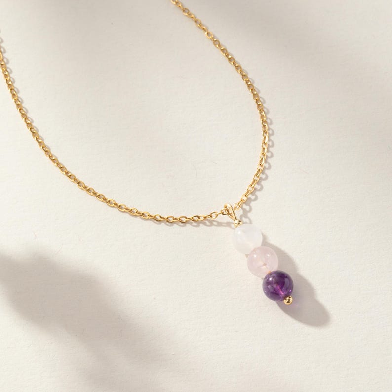 May include: A gold chain necklace featuring a vertical pendant composed of three round beads arranged in a gradient from white to light pink to deep purple. The pendant is attached to the delicate gold link chain with a small gold jump ring.