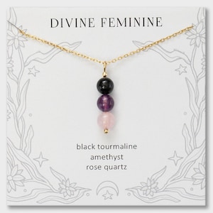 May include: A gold chain necklace featuring a vertical pendant with three round beads: a black tourmaline bead, a purple amethyst bead, and a pale pink rose quartz bead. The display card reads DIVINE FEMININE, black tourmaline, amethyst, rose quartz.