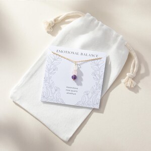 May include: A gold-tone chain necklace with three small beads in moonstone, rose quartz, and amethyst, displayed on a white card labeled EMOTIONAL BALANCE with floral line art, resting on a white fabric drawstring pouch with knotted ends.