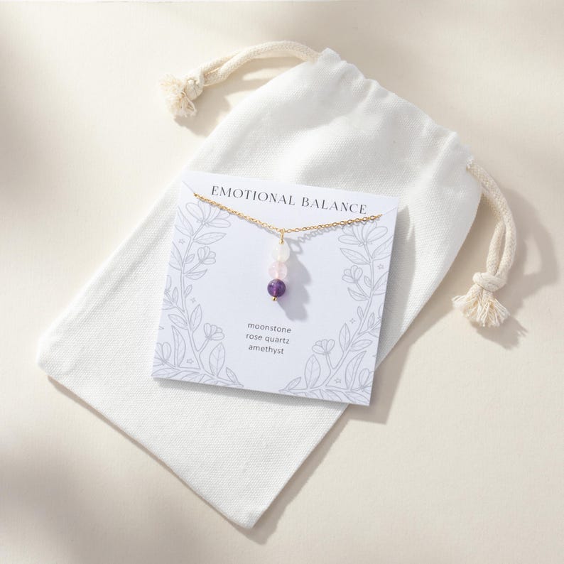 May include: A gold-tone chain necklace with three small beads in moonstone, rose quartz, and amethyst, displayed on a white card labeled EMOTIONAL BALANCE with floral line art, resting on a white fabric drawstring pouch with knotted ends.