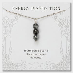 May include: A silver-toned chain necklace featuring a vertical pendant with three round beads: tourmalated quartz, black tourmaline, and hematite. The display card reads ENERGY PROTECTION, tourmalated quartz, black tourmaline, and hematite.