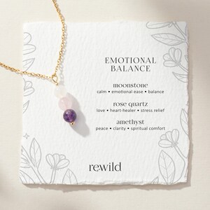 May include: A gold-tone chain necklace featuring three round beads: a white moonstone, a pale pink rose quartz, and a deep purple amethyst. The necklace is displayed on a square card with the text EMOTIONAL BALANCE and the brand name rewild.