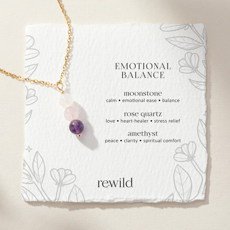 May include: A gold-tone chain necklace featuring three round beads: a white moonstone, a pale pink rose quartz, and a deep purple amethyst. The necklace is displayed on a square card with the text EMOTIONAL BALANCE and the brand name rewild.