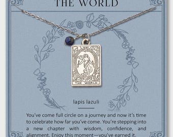 Può includere: Collana color argento con un ciondolo rettangolare con la scritta "THE WORLD" e un'illustrazione. È presente anche una piccola perla di lapislazzuli blu scuro. La collana è presentata su una carta blu con testo.