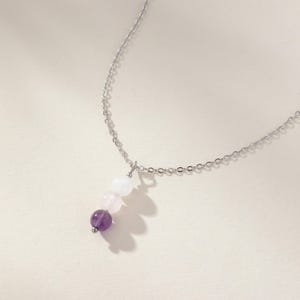 May include: A silver-toned metal chain necklace featuring a vertical pendant composed of three round beads. The beads are arranged in a gradient from top to bottom, starting with a translucent white bead, followed by a pale pink bead, and a deep purple bead.