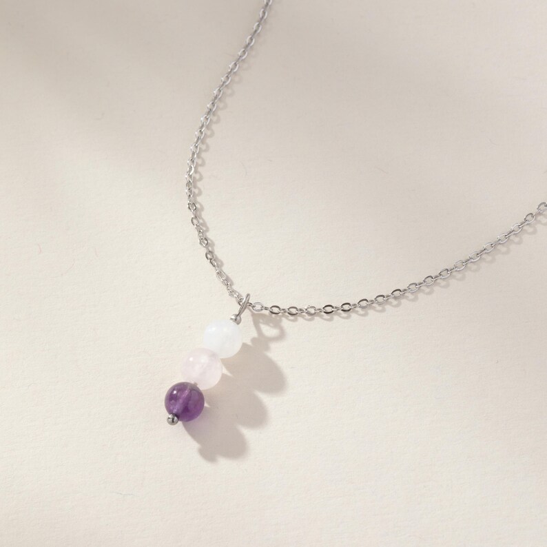 May include: A silver-toned metal chain necklace featuring a vertical pendant composed of three round beads. The beads are arranged in a gradient from top to bottom, starting with a translucent white bead, followed by a pale pink bead, and a deep purple bead.