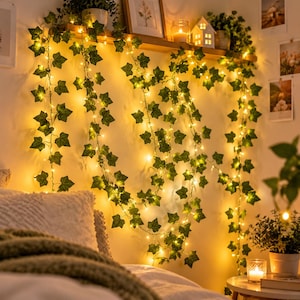 May include: A string of artificial green ivy leaves intertwined with warm white LED fairy lights. The vine features numerous small, pointed leaves and thin wire strands that emit a soft, glowing light, draped across a wall and shelf.