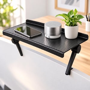 May include: A black carbon fiber textured monitor top shelf with two adjustable support legs. The shelf holds a smartphone, a cylindrical silver speaker, and a small green potted plant in a white ceramic pot, mounted on a white screen edge.