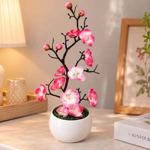 May include: A decorative artificial cherry blossom branch with vibrant pink and white petals and small buds, arranged on dark brown stems. The branch is planted in a round, smooth white ceramic pot filled with green moss at the base.