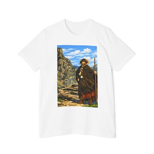 May include: A white short-sleeve t-shirt featuring a rectangular graphic print of a bearded figure wearing a dark hooded cloak with ram horns, holding a wooden staff while standing on a rocky mountain path under a bright blue sky with clouds.